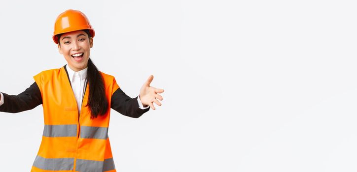 Friendly smiling asian female construction manager, engineer in safety helmet and reflective jacket, extending hands for greeting, welcome business partners, standing white background.
