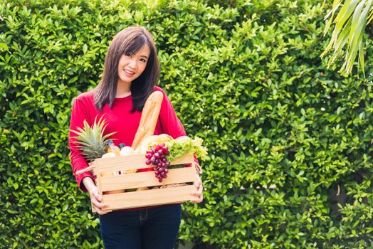 Portrait of Asian beautiful young woman farmer standing she smile and holding full fresh food raw vegetables fruit in a wood box in her hands on green leaves background