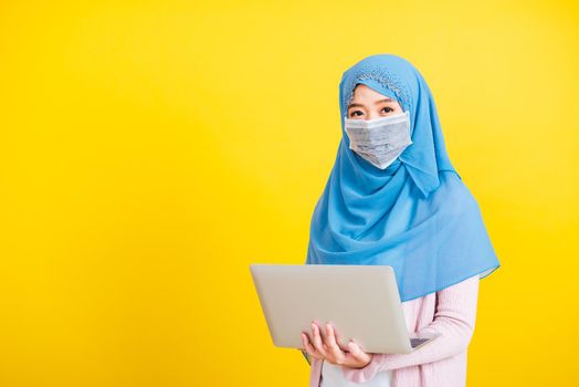 Asian Muslim Arab, Portrait of happy beautiful young woman religious wear veil hijab and face mask protective to prevent coronavirus she holding the laptop typing, isolated on yellow background