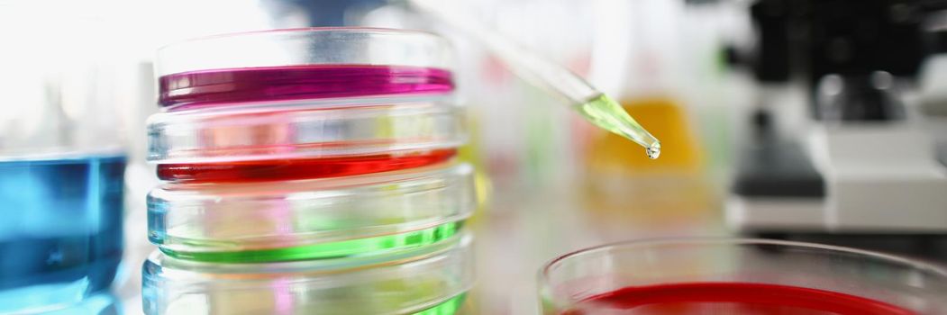 Close-up of lab worker adding drop of liquid into testing material with dropper. Colourful glass containers on surface. Research, experiment, science, discovery concept