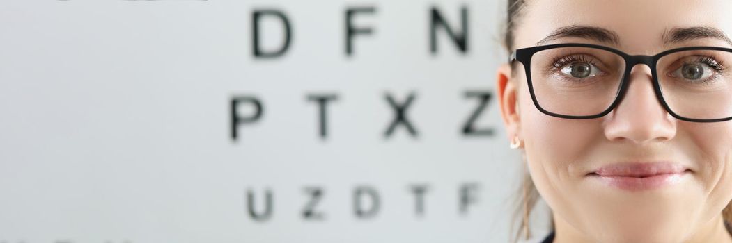 Portrait of eye doctor with eye test chart on background. Female optician in glasses, appointment in eye clinic. Copy space in left. Checkup, medicine, healthcare, ophthalmologist concept