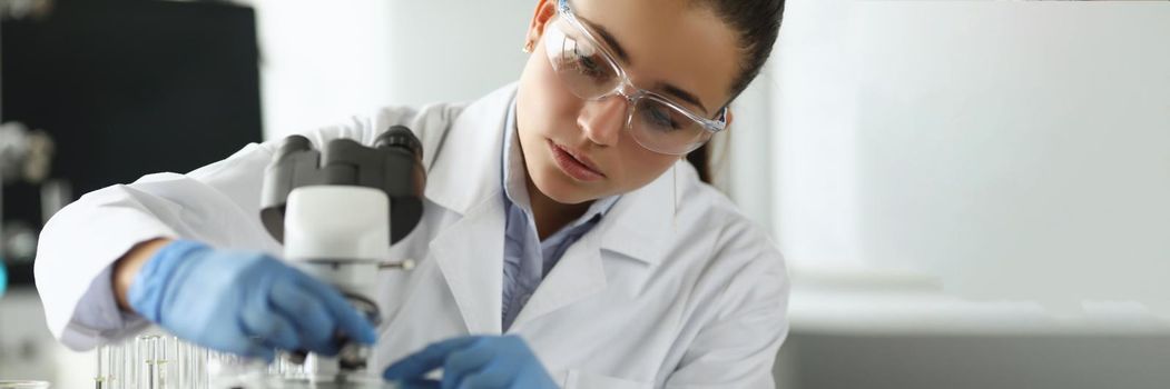 Portrait of smart young scientist performing experiment in laboratory. Concentrated biologist in uniform work with microscope in office. Science, investigation, discovery concept