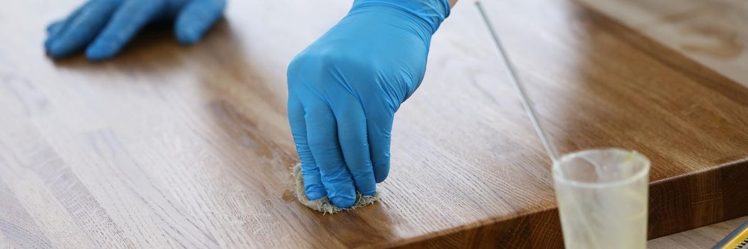 Close-up of worker hands in gloves polishing wood surface, cover table top with varnish. Tools for construction works on floor. Renovation, fix and repair furniture concept