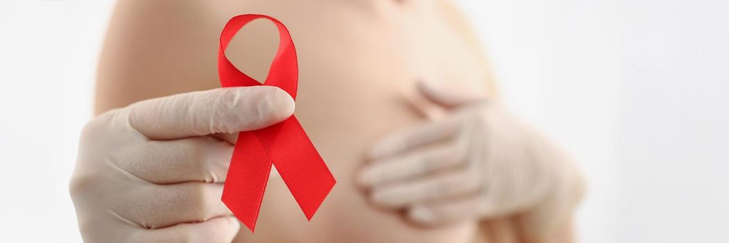 Close-up of woman holding red ribbon, fight breast cancer, support female in fighting illness. Topless female on blurred background. Breast cancer, disease, medicine, diagnostics concept