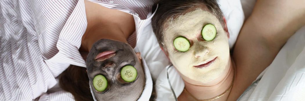 Top view of couple making beauty treatment together, hydrating face masks and cucumbers pieces on eyes. Spa day at home, lazy weekend, comfy pyjamas, wellness, skincare concept
