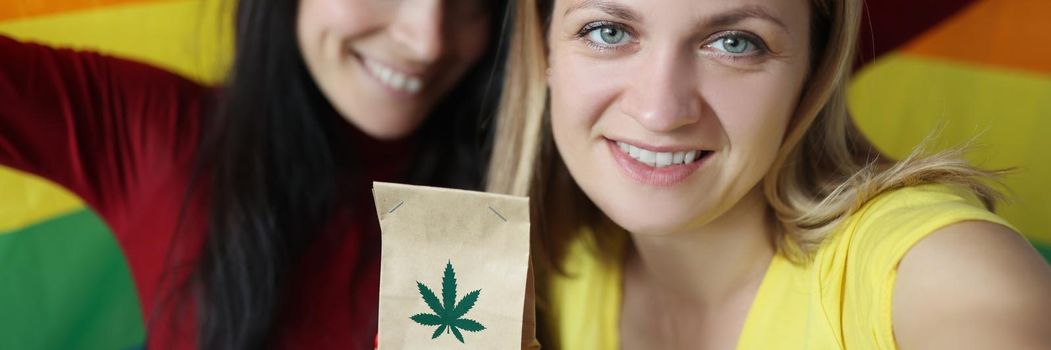 Portrait of brunette and blonde women hold package with hemp plant, paper package with marijuana. Treating pain, stress and insomnia. Legalized narcotic substance, fun concept