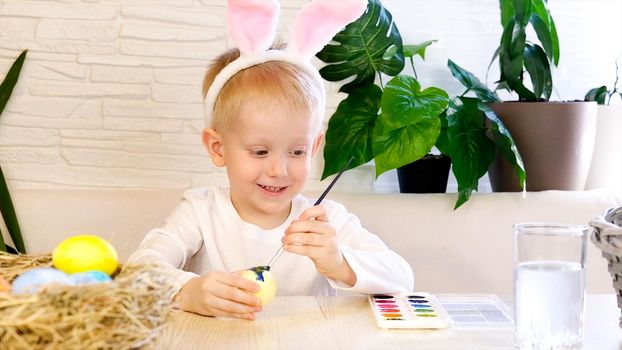 A little boy in rabbit ears enthusiastically paints Easter eggs with paint. Religious holidays concept, do it yourself, family leisure, easter egg hunt.