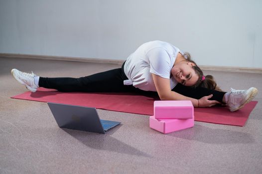 Young caucasian fat woman doing bends on a sports mat and watching a training video on a laptop. A chubby girl stretches the split remotely using video communication.