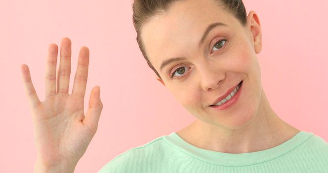 Friendly looking polite young Caucasian woman saying hi, waving her hand. Positive human emotions, feelings,facial expressions, attitude and reaction at pink background