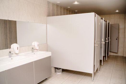 The interior of a public toilet. Row of wash basins with metal faucets on a marble slab, liquid soap dispensers, a long mirror on a gray wall.