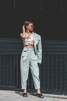 Fashion portrait of young woman wearing sunglasses, top, slingbacks, blue suit. Young beautiful happy model posing near gray metal grid.