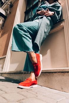 Woman in a red stylish suede loafer shoes, blue jeanse, white shirt and trendy checkered coat. Pair of trendy female loafers shoes, outdoors