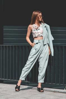 Fashion portrait of young woman wearing sunglasses, top, slingbacks, blue suit. Young beautiful happy model posing near gray metal grid.