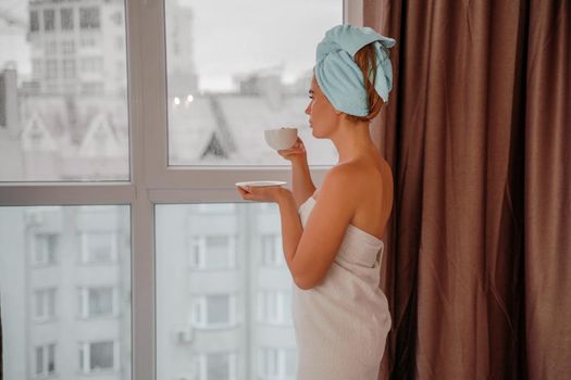 Young serene relaxed woman in spa bath towel drinking hot beverage tea coffee after taking shower bath at home. Beauty treatment, hydration concept