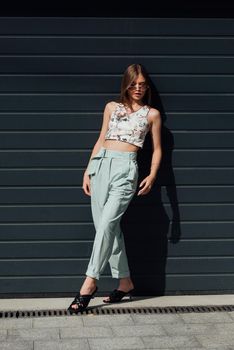 Fashion portrait of young woman wearing sunglasses, top, slingbacks, blue suit. Young beautiful happy model posing near gray metal grid.