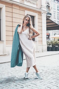 Fashionable young woman with blond hair dressed in beige dress, checkered coat and sneakers posing. Street style.