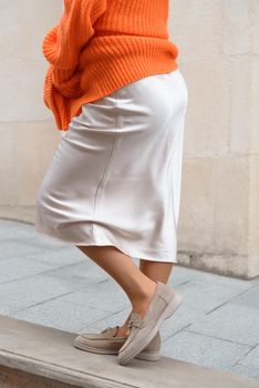 Portrait of fashionable blond women in orange sweater, beige dress and stylish suede loafer shoes posing in the street