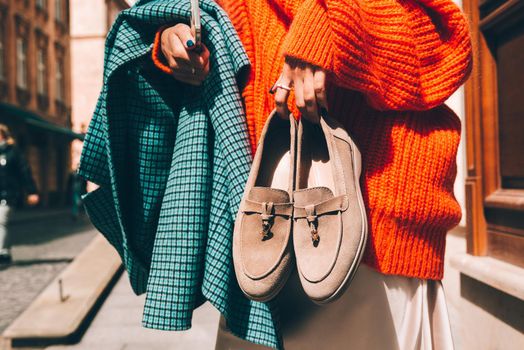 Close-up photo of fashionable women in orange sweater and beige dress holding in hands checkered coat and beige suede shoes. No face