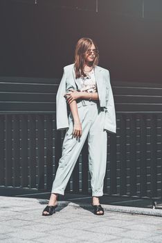 Fashion portrait of young woman wearing sunglasses, top, slingbacks, blue suit. Young beautiful happy model posing near gray metal grid.