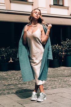 Fashionable young woman with blond hair dressed in beige dress, checkered coat and sneakers posing. Street style.