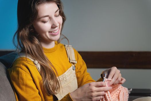 Young happy caucasian woman smiling sitting on sofa and crocheting wool product. Women's hobby production of clothes from wool.
