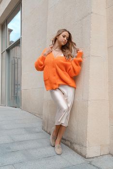 Portrait of fashionable blond women in orange sweater, beige dress and stylish suede loafer shoes posing in the street