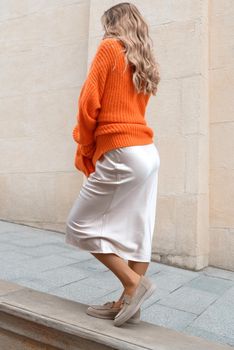 Portrait of fashionable blond women in orange sweater, beige dress and stylish suede loafer shoes posing in the street