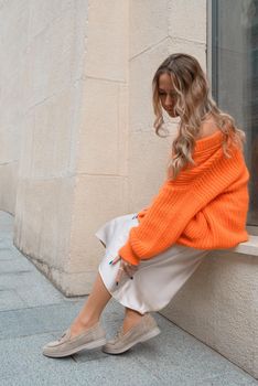 Portrait of fashionable blond women in orange sweater, beige dress and stylish suede loafer shoes posing in the street