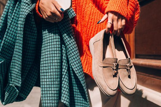 Close-up photo of fashionable women in orange sweater and beige dress holding in hands checkered coat and beige suede shoes. No face