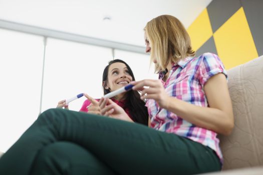 Low angle of friends girls hold pregnancy tests, happy best friends got big news. Smiling women on sofa at home. Friendship, pregnancy, new period, life change, happiness concept