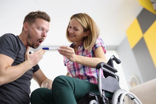 Low angle of woman in wheelchair show to husband positive pregnancy test. Surprised reaction of man, bright emotion. Disabled people, family growing, happiness, healthcare concept