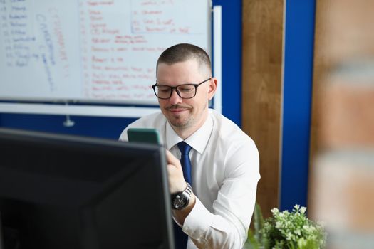 Portrait of company boss texting in smartphone, leader in private office. Computer on workplace, board with lettering on wall. Business, working day, career growth, technology concept