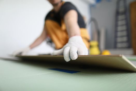 Close-up of carpenter laying wooden panel of laminate floor, installing new floor for apartment. Qualified worker deal with parquet pieces. Home renovation, construction concept. Blurred background