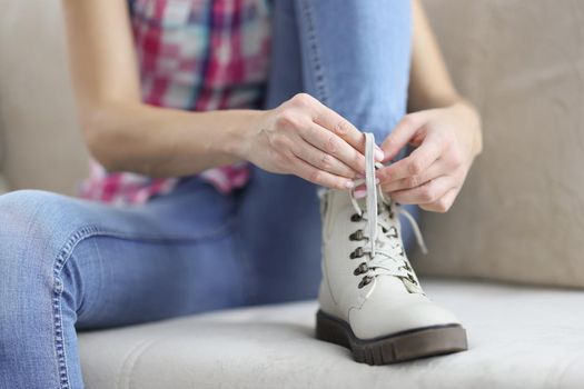 Close-up of woman sit on couch and tie shoelaces on boots, female placed foot on clean sofa. Fix shoes before going out. New footwear, purchase, shopping, fashion, style concept