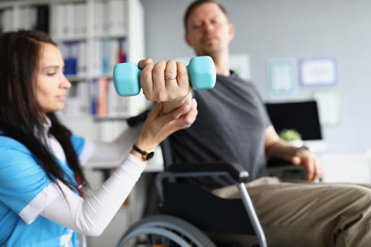 Close-up of physiotherapist woman doing treatment with patient in wheelchair. Exercise with dumbbell equipment for arm function. Medicine, clinic, recovery, healthcare, injury concept