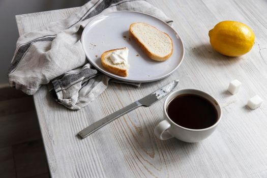 A cup of coffee, two pieces of sugar, a lemon, a piece of bread with curd cheese spread on it on a white ceramic plate with a knife on the table.