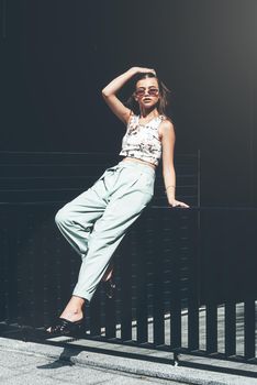 Fashion portrait of young woman wearing sunglasses, top, slingbacks, blue suit. Young beautiful happy model posing near gray metal grid.