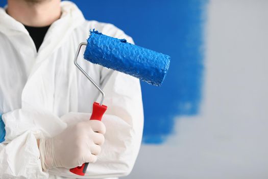 Close-up of painter man hand holding roller tool for painting walls, blue colour on equipment, worker in uniform. Copy space in right. Renovation, interior design, redecoration concept