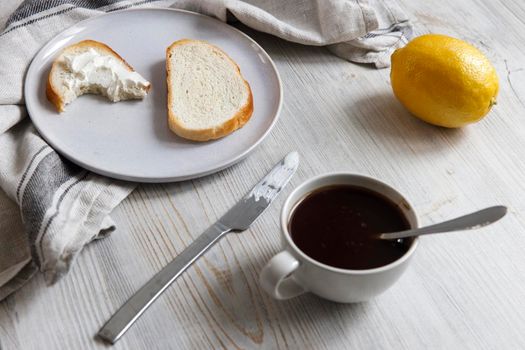 A cup of coffee, two pieces of sugar, a lemon, a piece of bread with curd cheese spread on it on a white ceramic plate with a knife on the table.