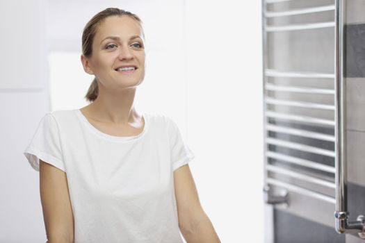 Portrait of young woman looking in mirror at herself in bathroom, white smile, natural beauty, no makeup, morning routine, light restroom. Good morning, wellness, beauty, youth concept