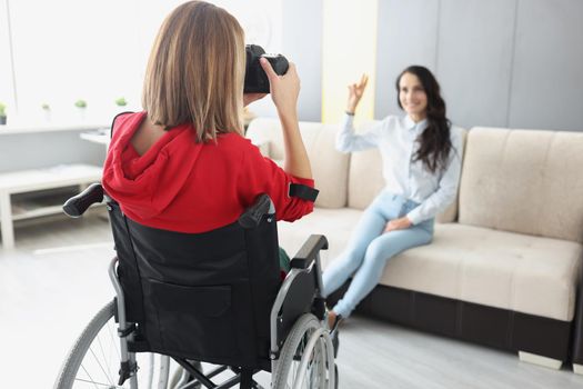 Close-up of woman photographer in wheelchair taking picture of client on sofa, hobby as job, creative unique photoshot for friend. Art, creativity, disabled people concept. Blurred background