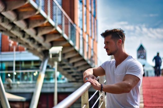 Handsome fit man in white t-shirt outdoor in city setting, leaning against handrail, looking away