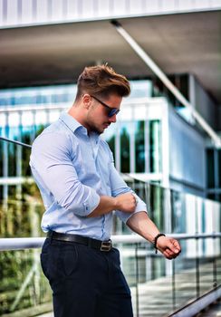 One handsome elegant man in urban setting in European city, standing, rolling up shirt's sleeves