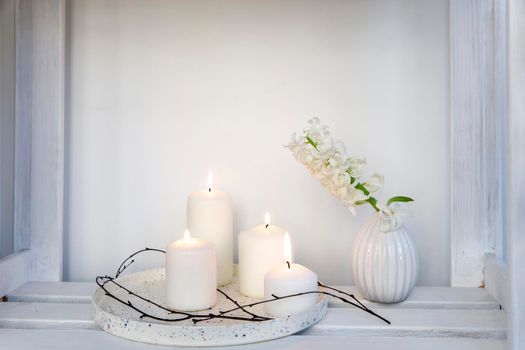 A bouquet of white cut hyacinth a in a small white corrugated vase and three large burning candles on a round tray are on a beige table. Place for text