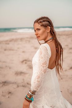 Model in boho style in a white long dress and silver jewelry on the beach. Her hair is braided, and there are many bracelets on her arms