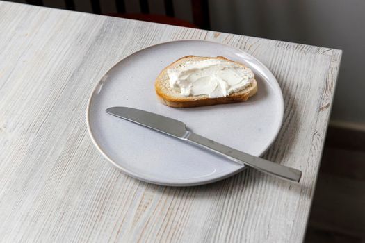 A piece of bread with curd cheese spread on it on a white ceramic plate with a knife on the beige table.