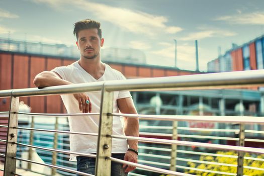 Handsome fit man in white t-shirt outdoor in city setting, leaning against handrail, looking away