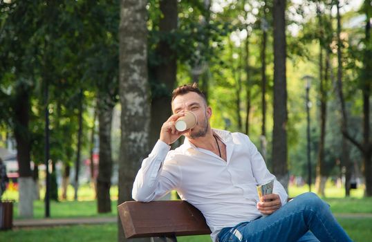 A happy man sits on a bench, drinks coffee, holds dollars. A young man on a background of green trees, a hot sunny summer day. Warm soft light, close-up.