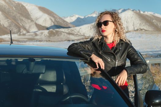 Portrait of a stylish attractive Caucasian young woman getting out of a retro sports car in the mountains at sunset. Travel concept copy space.
