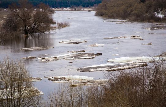 White ice floes float down the river slowly. Spring, snow melts, dry grass all around, floods begin and the river overflows. Day, cloudy weather, soft warm light.